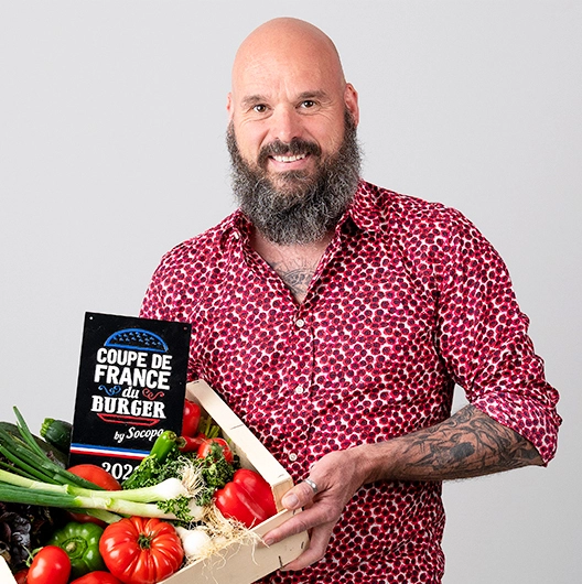 Sophie-mare-Photographe-seingbouse-portrait-corporate-professionel-profil-cv-linkedin-site-internet-01 restaurateur tatoué chauve et barbu avec chemise à motifs rouges, avec une caisse de légumes et la pancarte de coupe de france du burger