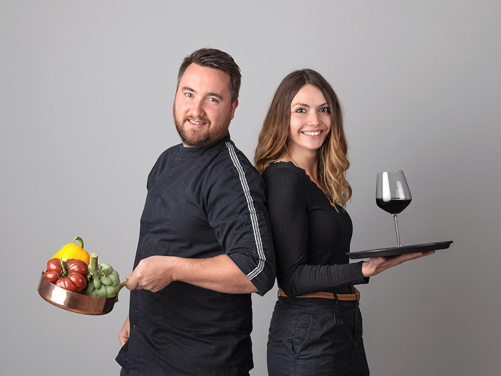 Sophie-mare-Photographe-seingbouse-portrait-corporate-professionel-profil-cv-linkedin-site-internet-03 couple homme et femme dos à dos en tenues noires, la femme tient un plateau sur sa main avec un verre de vin rouge et l'homme tient une casserole en cuivre avec des légumes