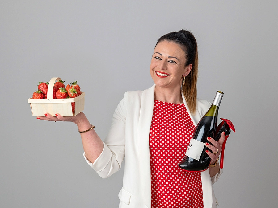 Sophie-mare-Photographe-seingbouse-portrait-corporate-professionel-profil-cv-linkedin-site-internet-11 femme souriante en veste blanche et pull rouge à pois blanc, elle tient une caisse de fraises et une bouteille de vin rouge