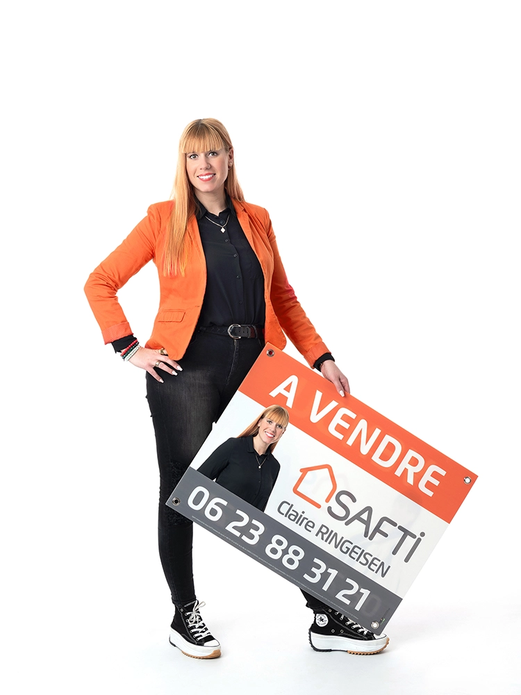 Sophie-mare-Photographe-seingbouse-portrait-corporate-professionel-profil-cv-linkedin-site-internet-19 une femme agent immobilier avec une veste orange et une tenue noire, debout sur fond blanc, avec un panneau à vendre en main
