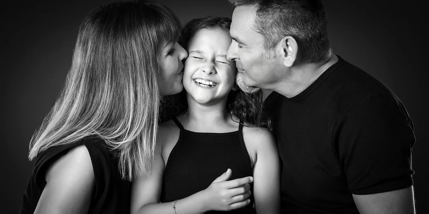 petite fille qui reçoit un bisou de son papa et sa maman en tenue noire sur fond noir