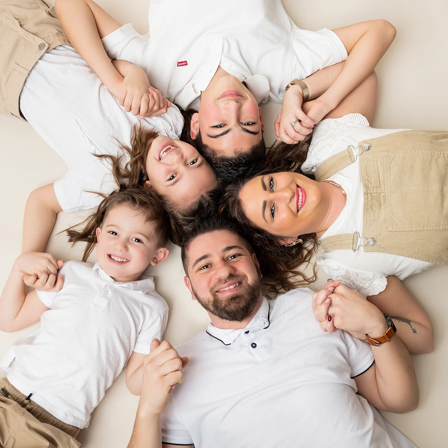 une famille de 5 personnes papa maman et 3 enfants, couchés au sol, mains tenues, sur fond beige