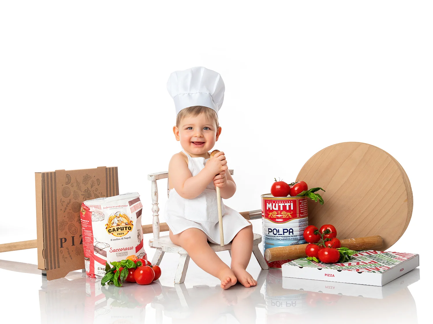 studio-seingbouse-photographe-sophie-julien-mare-fratrie-enfant-parents-frere-soeur-portrait-18 petit garçon d'1 an assis sur une chaise blanche avec une décoration de pizzaiolo, farine, tomates, sauces tomates, boite à pizza sur fond blanc