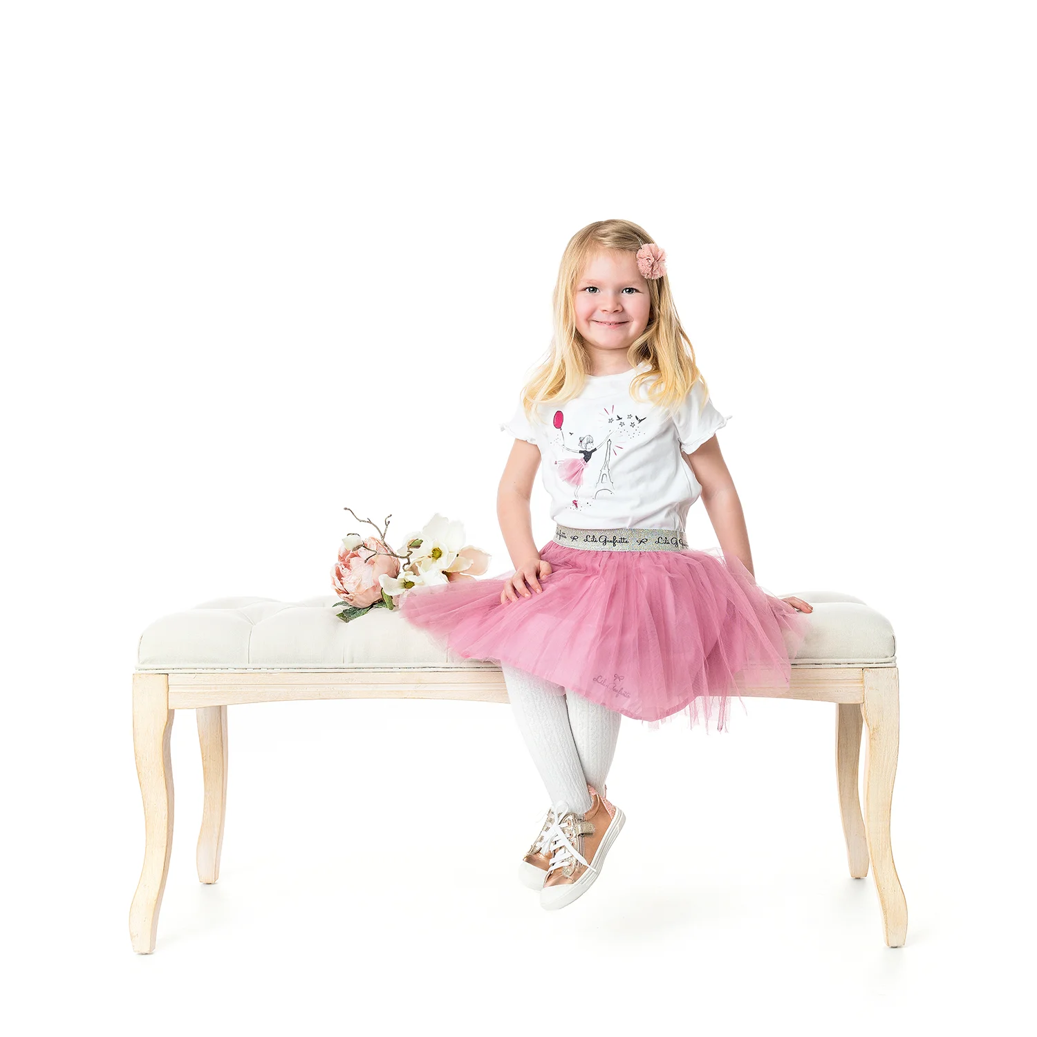 studio-seingbouse-photographe-sophie-julien-mare-fratrie-enfant-parents-frere-soeur-portrait-30 petite fille blonde souriante, assise sur un banc beige, avec des fleurs, sur fond blanc