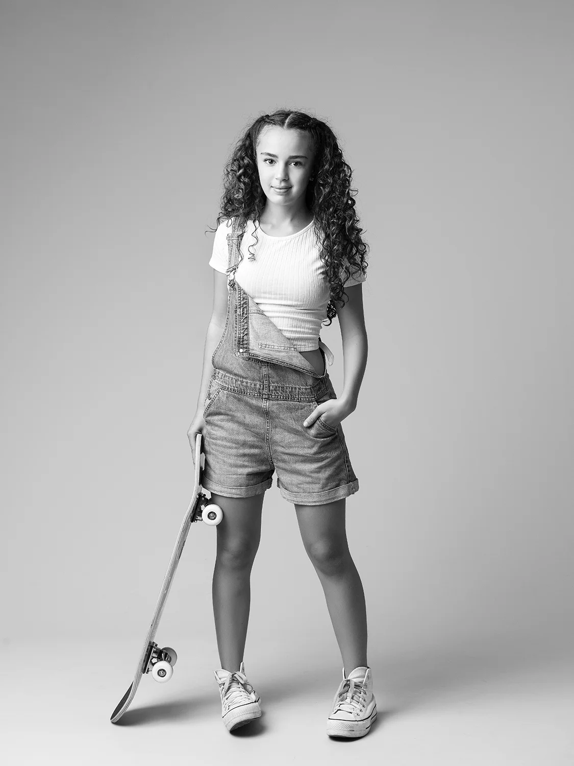 studio-seingbouse-photographe-sophie-julien-mare-fratrie-enfant-parents-frere-soeur-portrait-39 jeune ado cheveux longs bouclés en salopette en jean ouverte d'un coté avec un skate board debout sur fond gris