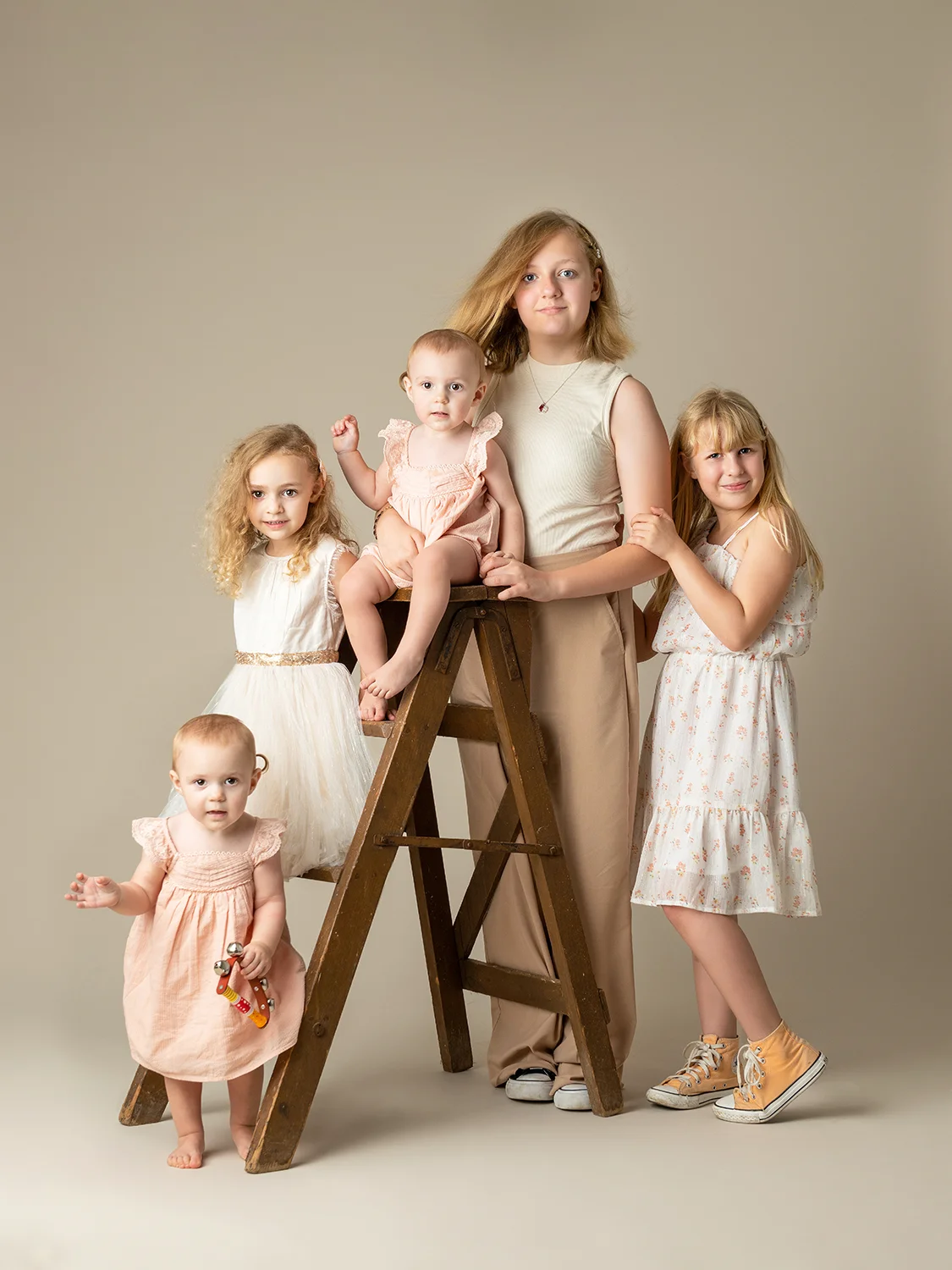 studio-seingbouse-photographe-sophie-julien-mare-fratrie-enfant-parents-frere-soeur-portrait-45 5 soeurs en tenues beiges avec une échelle en bois sur fond beige, avec des jumelles d'1 an
