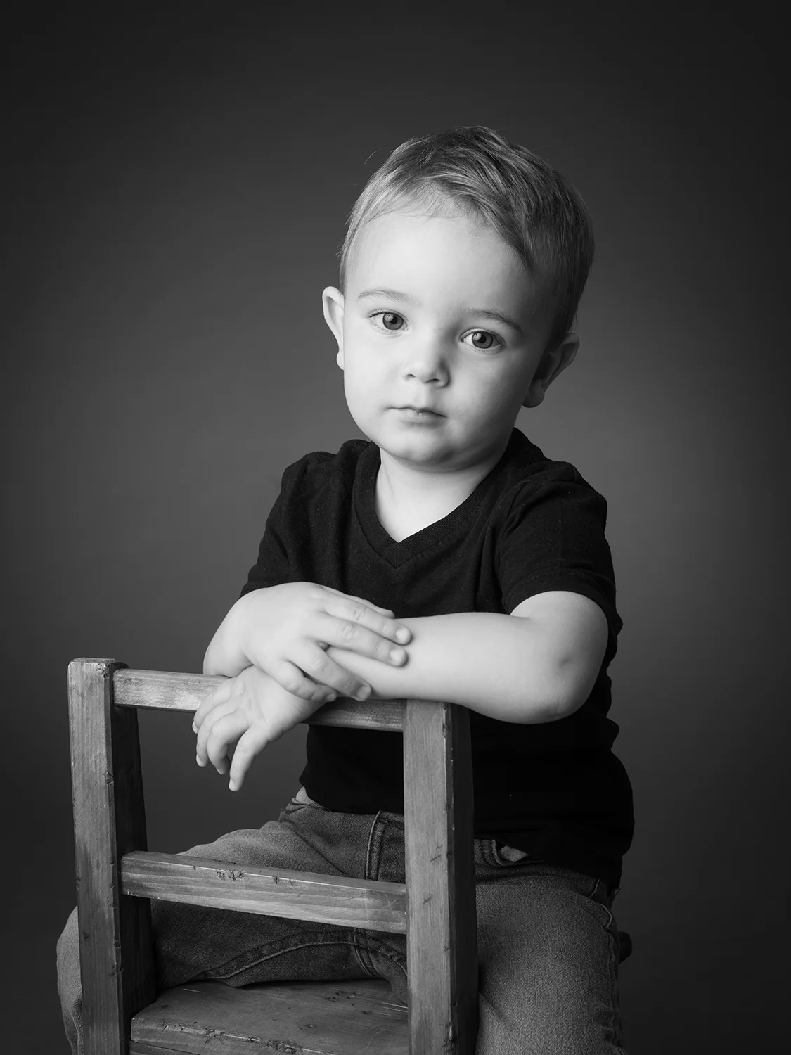 studio-seingbouse-photographe-sophie-julien-mare-fratrie-enfant-parents-frere-soeur-portrait-48 petit garçon avec un air triste assis sur une chaise en boir qui regarde l'objectif sur fond noir