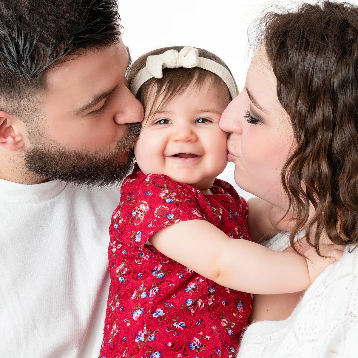 studio-seingbouse-photographe-sophie-julien-mare-fratrie-enfant-parents-frere-soeur-portrait-55 petite fille bébé qvec un bandeau et une robe rouge qui reçoit un bisou de son papa et sa maman et qui sourit