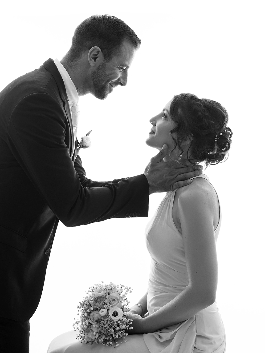 studio-sophie-mare-mariage-photos-seingbouse-merlebach-portrait-15 la mariée est assise, sur fond blanc. le marié la tient au cou et lui sourit. ils se regardent.