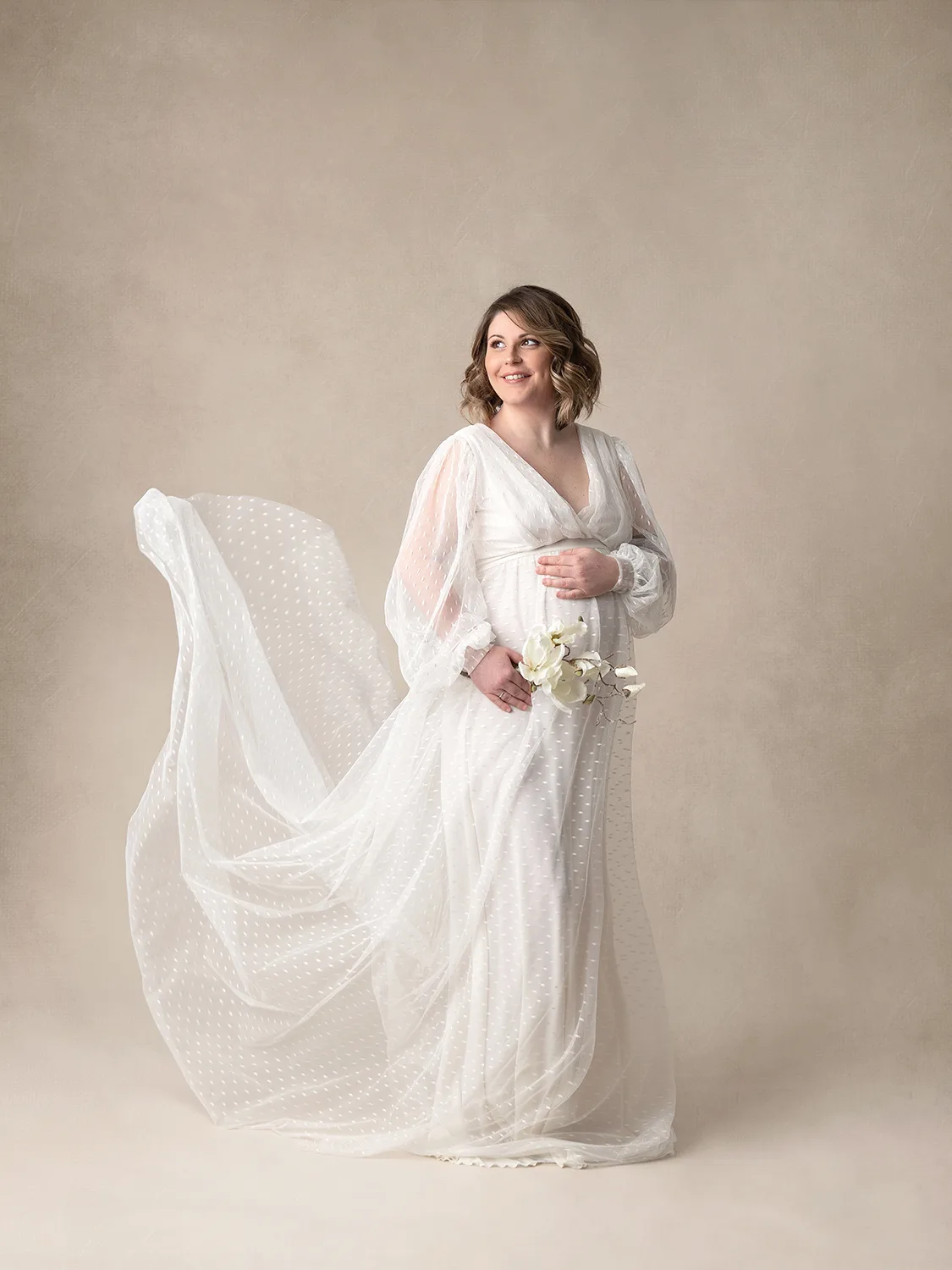 studio-seingbouse-photographe-sophie-julien-mare-grossesse-enceinte-parents-bebe-naissance-shooting-01 femme enceinte souriante, dans une robe beige qui vole, avec un fond beige, avec des fleurs dans la main
