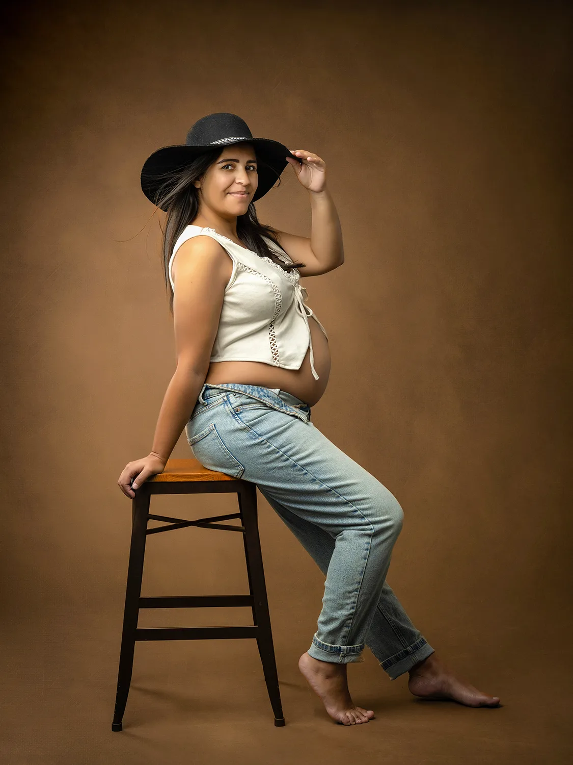 studio-seingbouse-photographe-sophie-julien-mare-grossesse-enceinte-parents-bebe-naissance-shooting-04 femme enceinte assise sur un tabouret, avec un jean et un chapeau noir