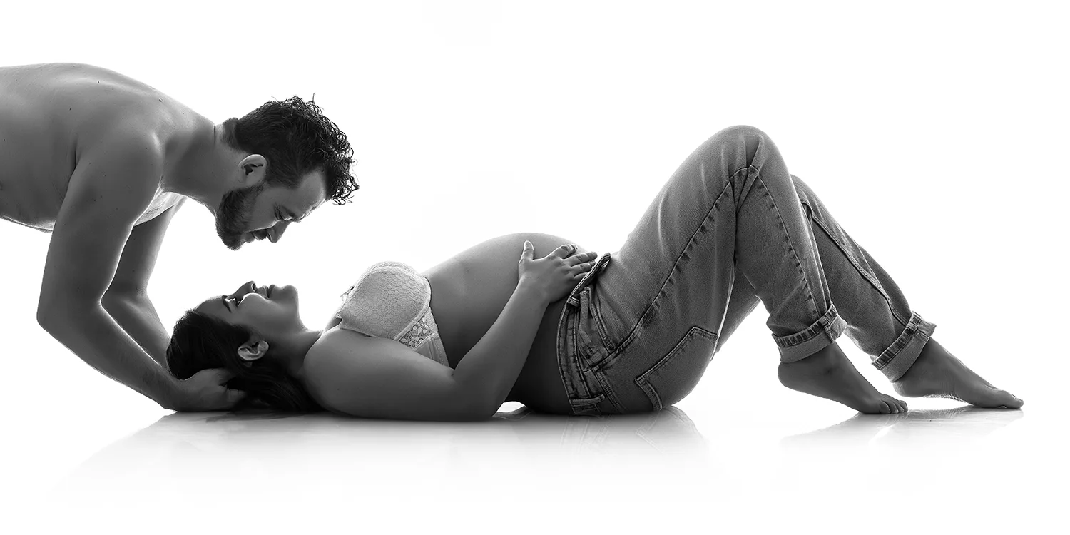 studio-seingbouse-photographe-sophie-julien-mare-grossesse-enceinte-parents-bebe-naissance-shooting-05 femme enceinte couchée sur le dos au sol, en jean et soutien-gorge blanc, son mari est au dessus d'elle