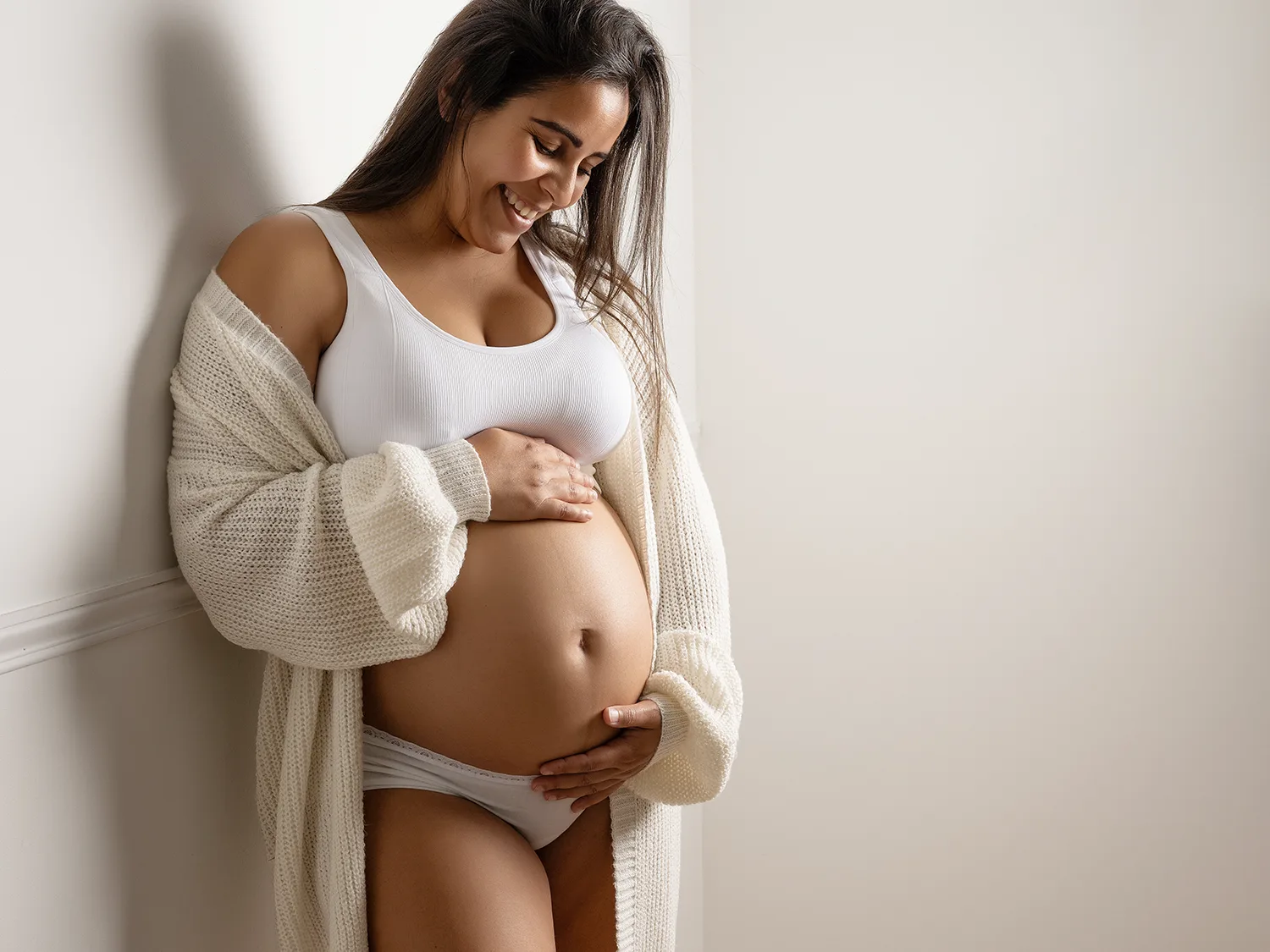 studio-seingbouse-photographe-sophie-julien-mare-grossesse-enceinte-parents-bebe-naissance-shooting-10 une future maman contre un mur beige, avec un gilet en laine, souriante