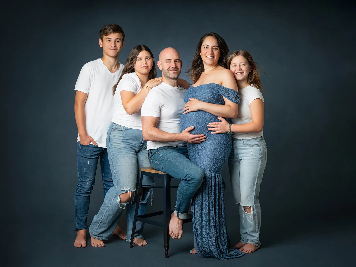 studio-seingbouse-photographe-sophie-julien-mare-grossesse-enceinte-parents-bebe-naissance-shooting-18 une famille de 5 personnes, la maman est enceinte et en robe bleue, les 4 autres personnes sont en jean et haut blanc, le papa est assis sur un tabouret
