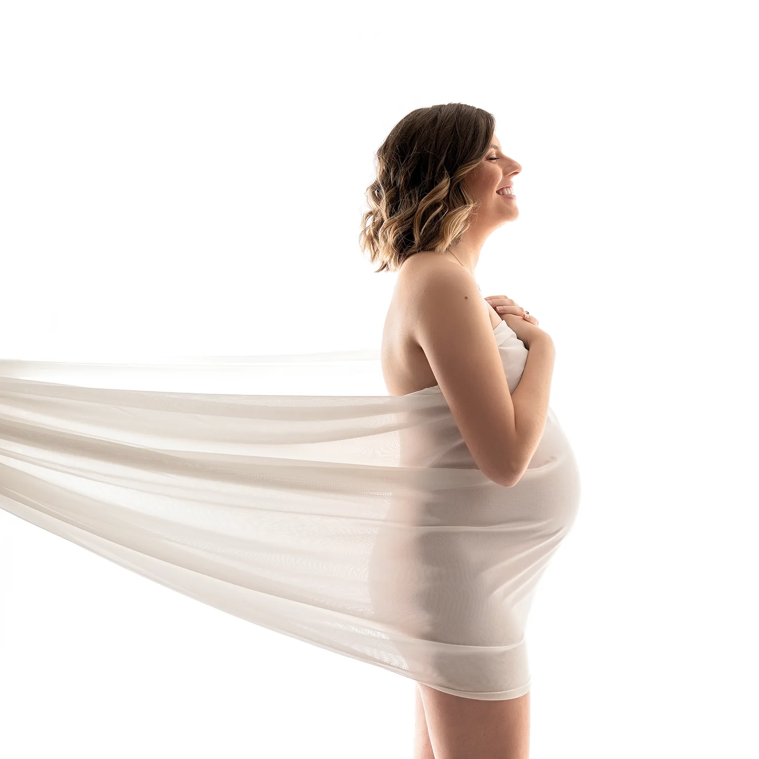 studio-seingbouse-photographe-sophie-julien-mare-grossesse-enceinte-parents-bebe-naissance-shooting-21 une femme enceinte souriante, de profil, sur fond blanc, entourée d'un voile blanc